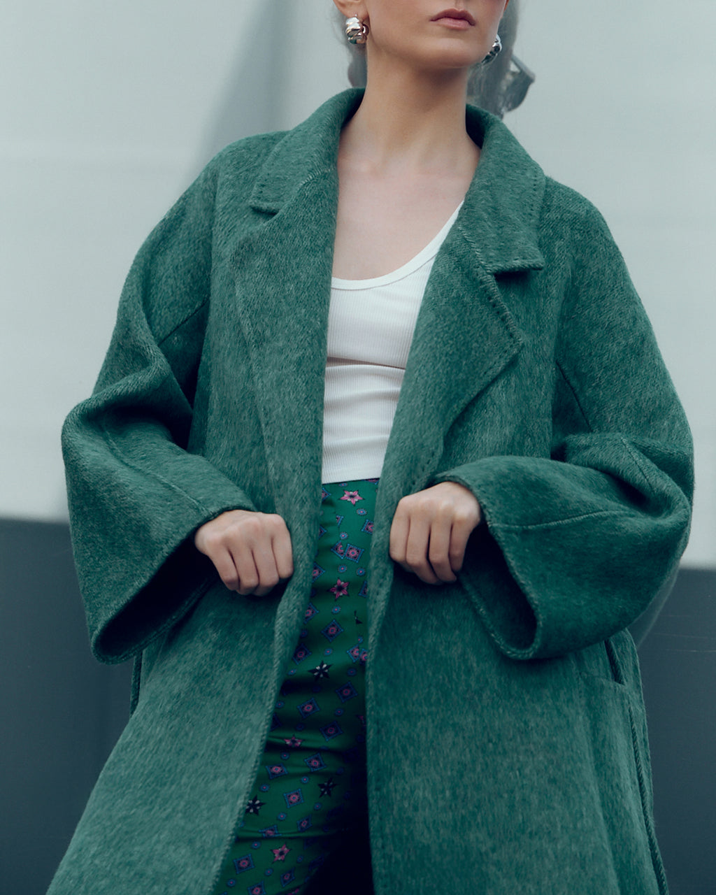 Long belted wool-blend coat in muted moss green, featuring wide lapels, oversized patch pockets, and a relaxed, robe-style silhouette. Made from a soft brushed wool and cashmere blend with a heathered texture, this timeless overcoat drapes elegantly for effortless city styling.