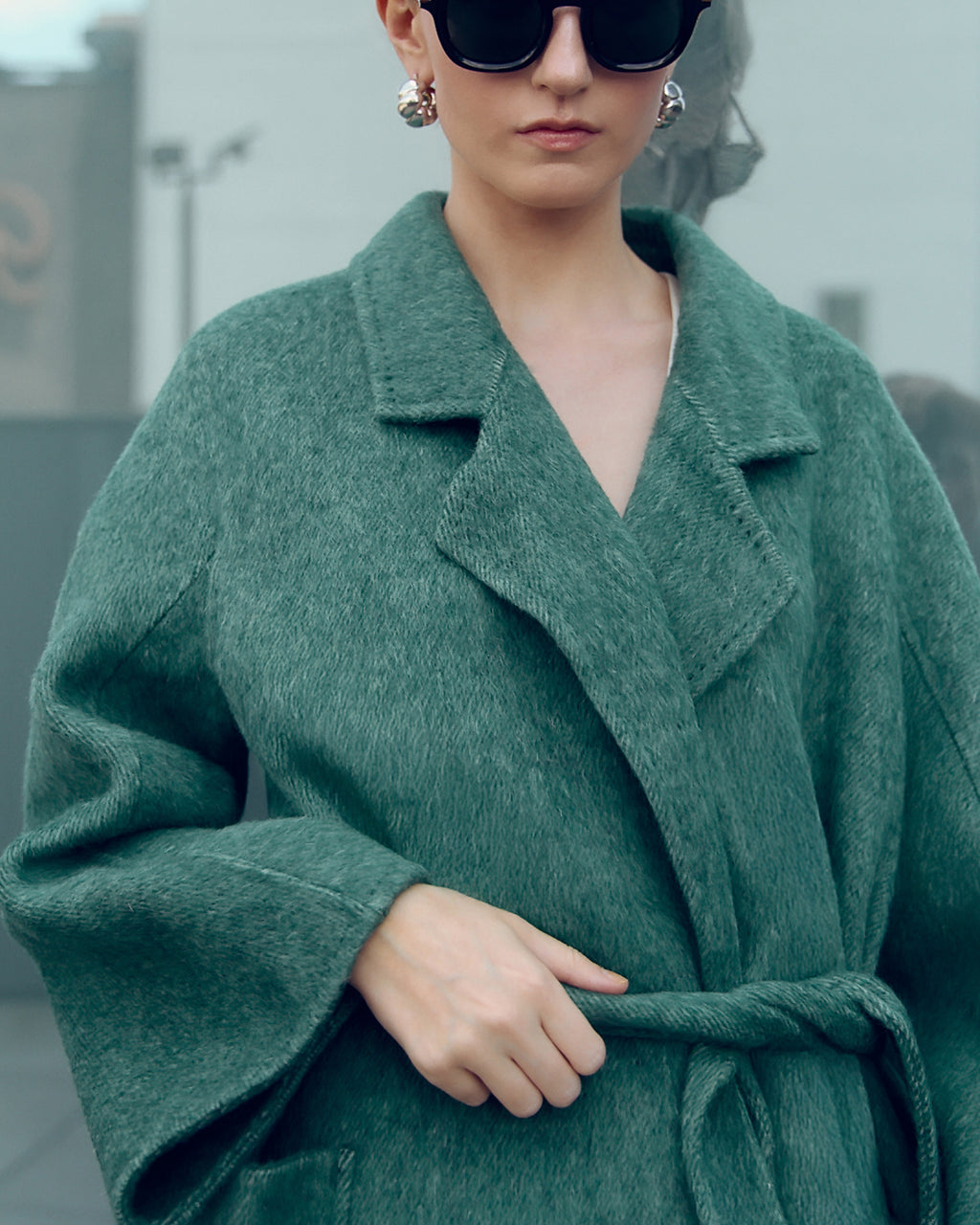 Long belted wool-blend coat in muted moss green, featuring wide lapels, oversized patch pockets, and a relaxed, robe-style silhouette. Made from a soft brushed wool and cashmere blend with a heathered texture, this timeless overcoat drapes elegantly for effortless city styling.