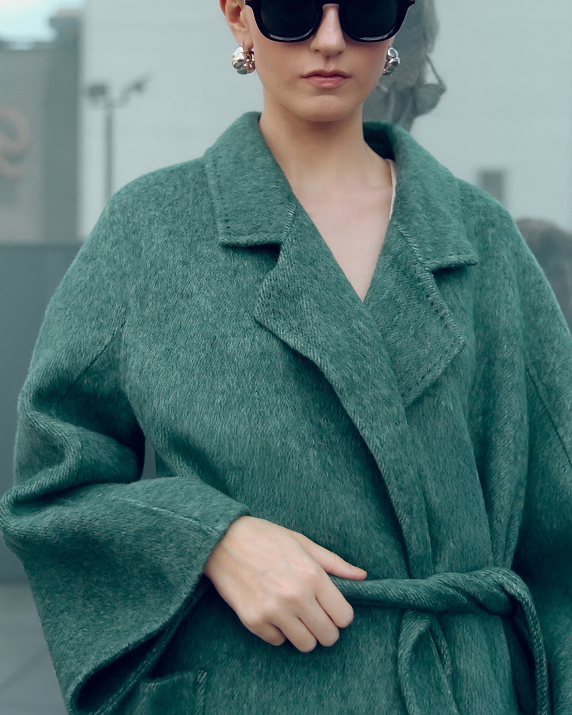 Long belted wool-blend coat in muted moss green, featuring wide lapels, oversized patch pockets, and a relaxed, robe-style silhouette. Made from a soft brushed wool and cashmere blend with a heathered texture, this timeless overcoat drapes elegantly for effortless city styling.