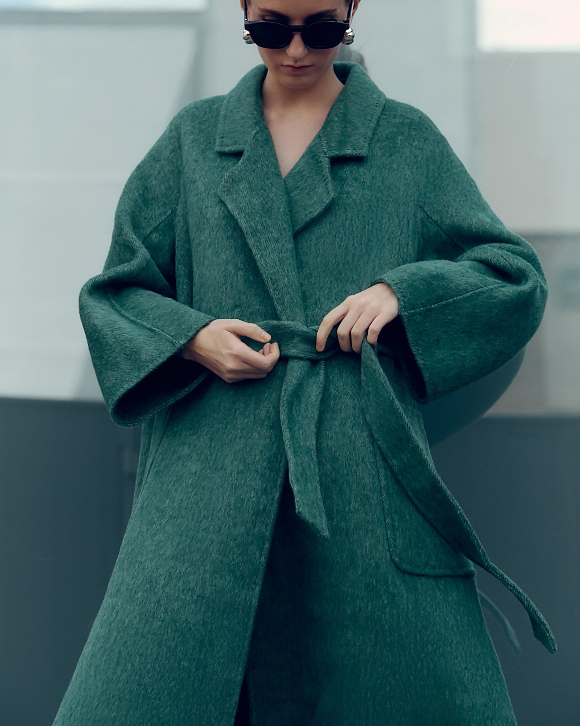 Long belted wool-blend coat in muted moss green, featuring wide lapels, oversized patch pockets, and a relaxed, robe-style silhouette. Made from a soft brushed wool and cashmere blend with a heathered texture, this timeless overcoat drapes elegantly for effortless city styling.