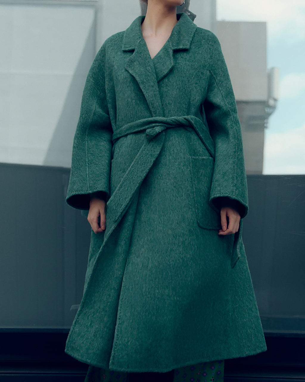 Long belted wool-blend coat in muted moss green, featuring wide lapels, oversized patch pockets, and a relaxed, robe-style silhouette. Made from a soft brushed wool and cashmere blend with a heathered texture, this timeless overcoat drapes elegantly for effortless city styling.